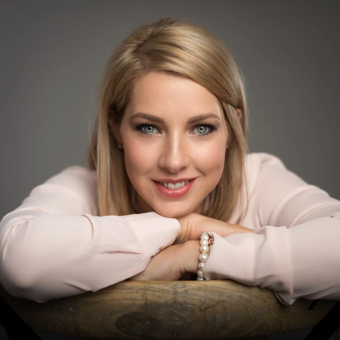 A smiling woman in a pink shirt leaning on the back of a chair