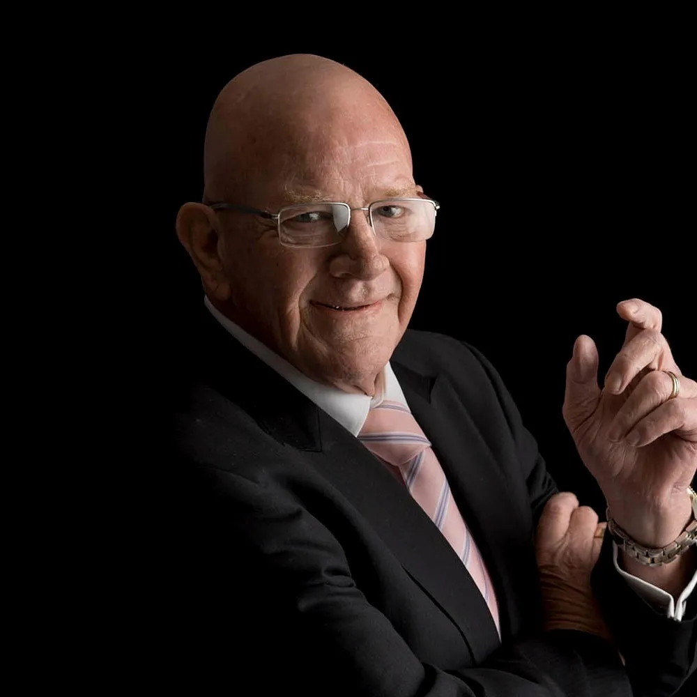 An older man in glasses and a dark suit against a black background