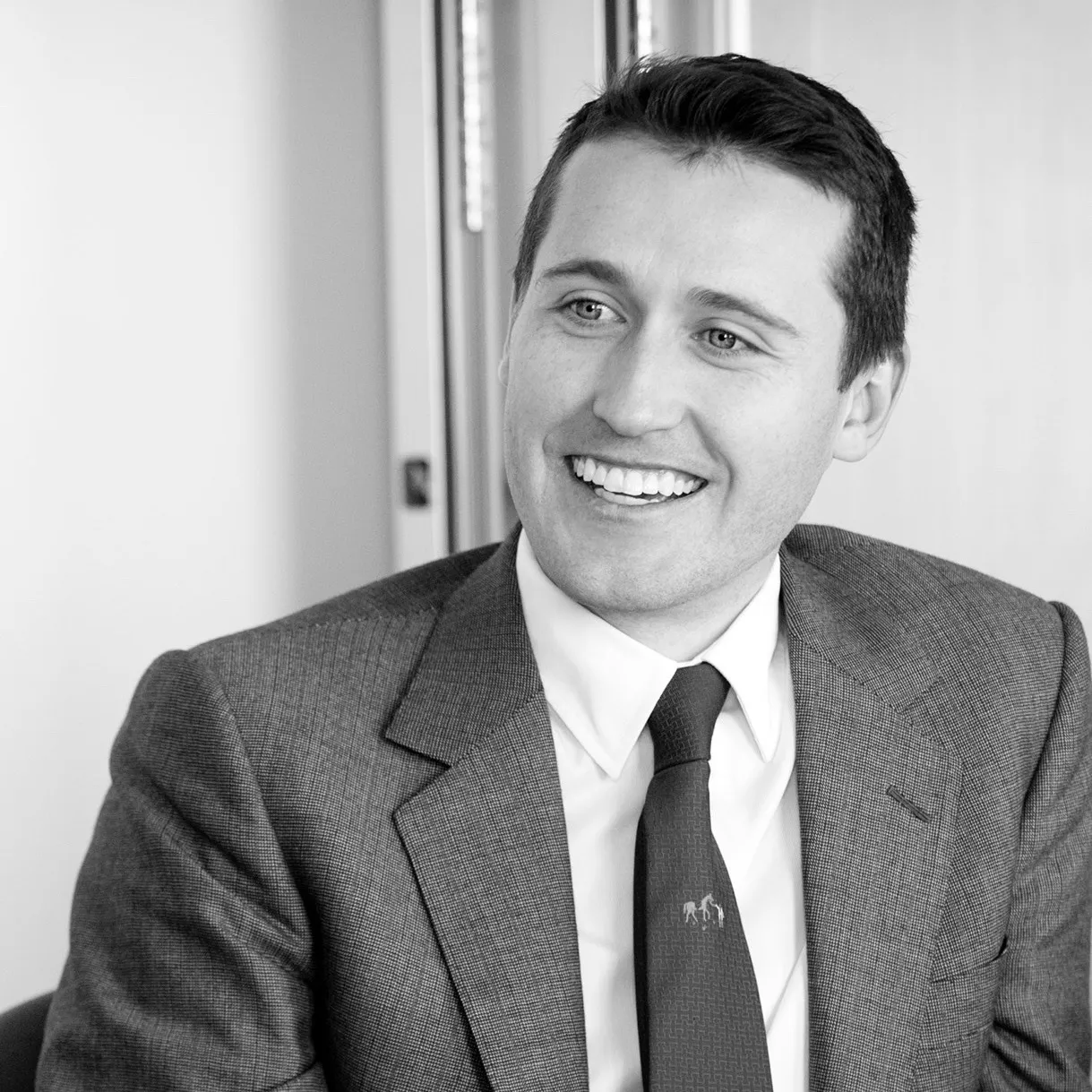 A smiling man in a grey suit and tie beside an office window, in black and white