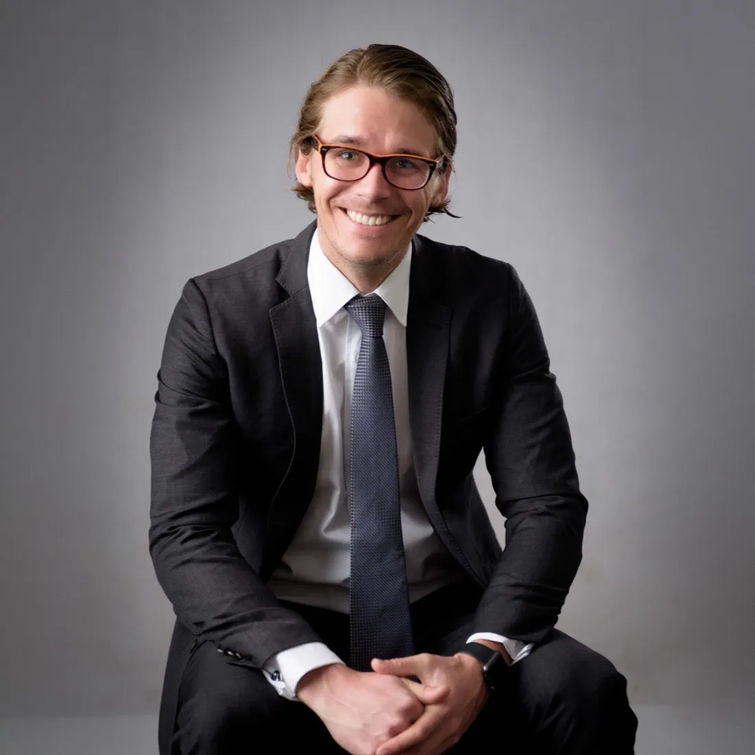 A young man in glasses and a dark suit, seated and smiling