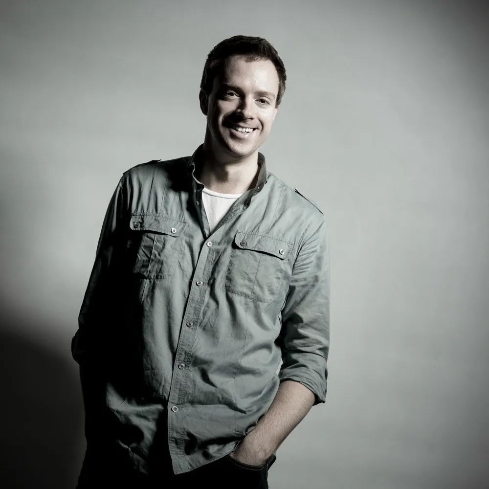 A smiling man in a casual shirt and jacket against a grey background