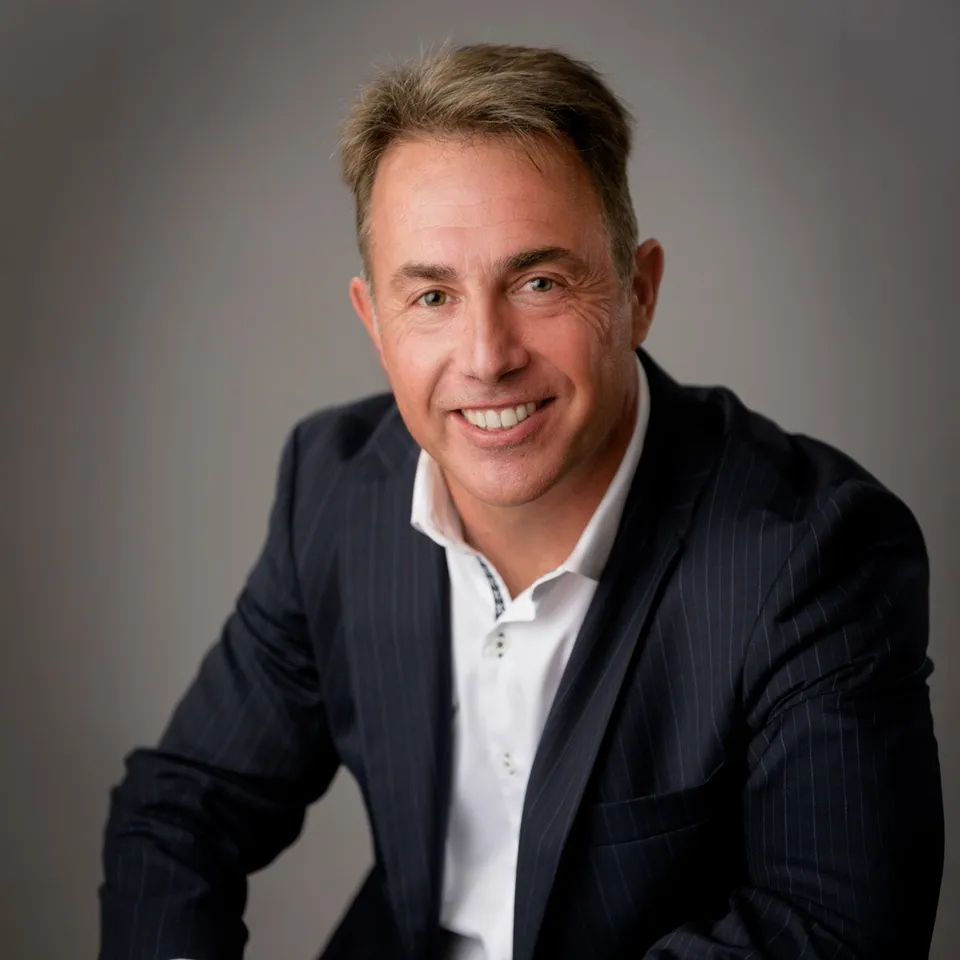 A smiling man in a navy jacket and open-collared white shirt
