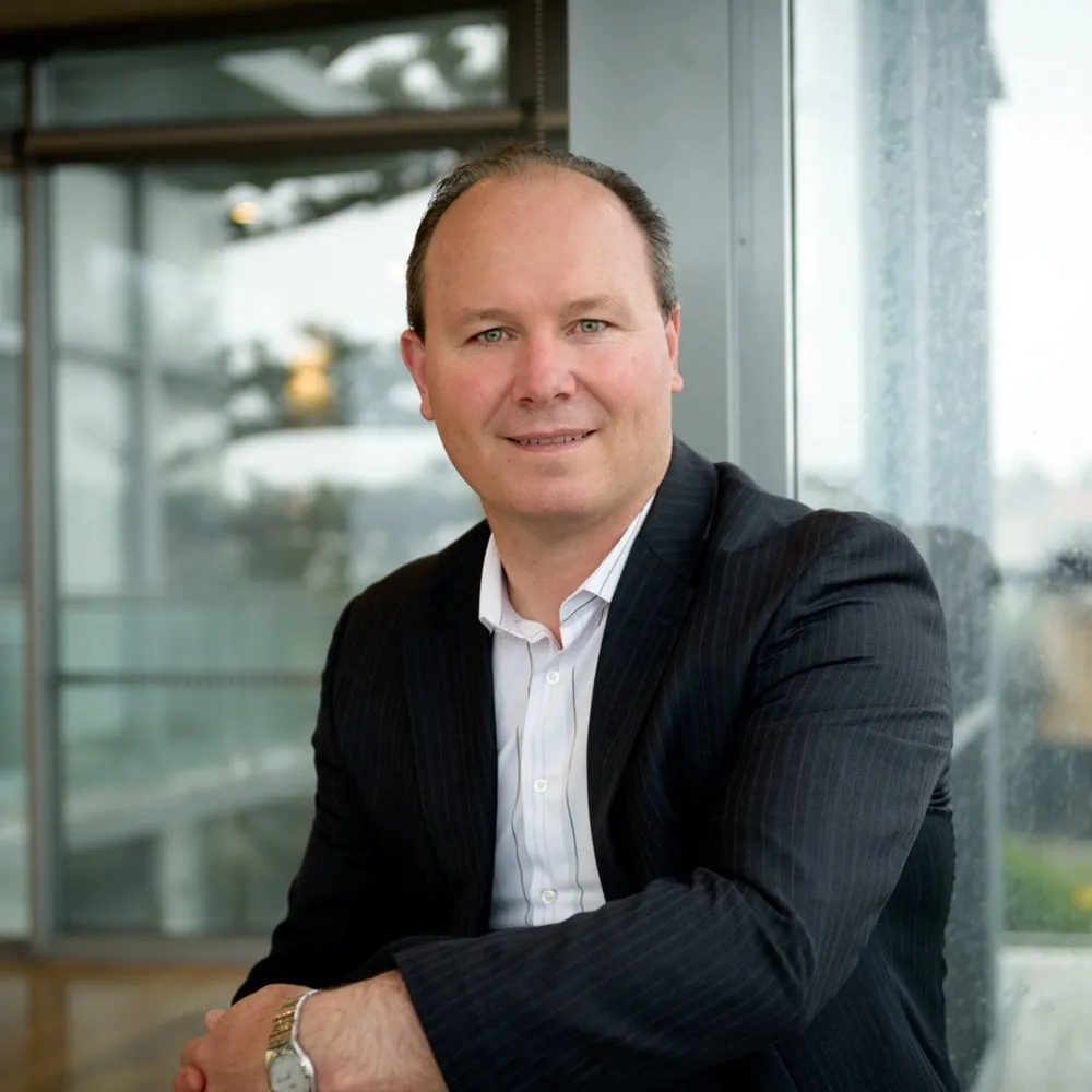 A smiling man in a dark jacket and open-collared shirt beside an office window