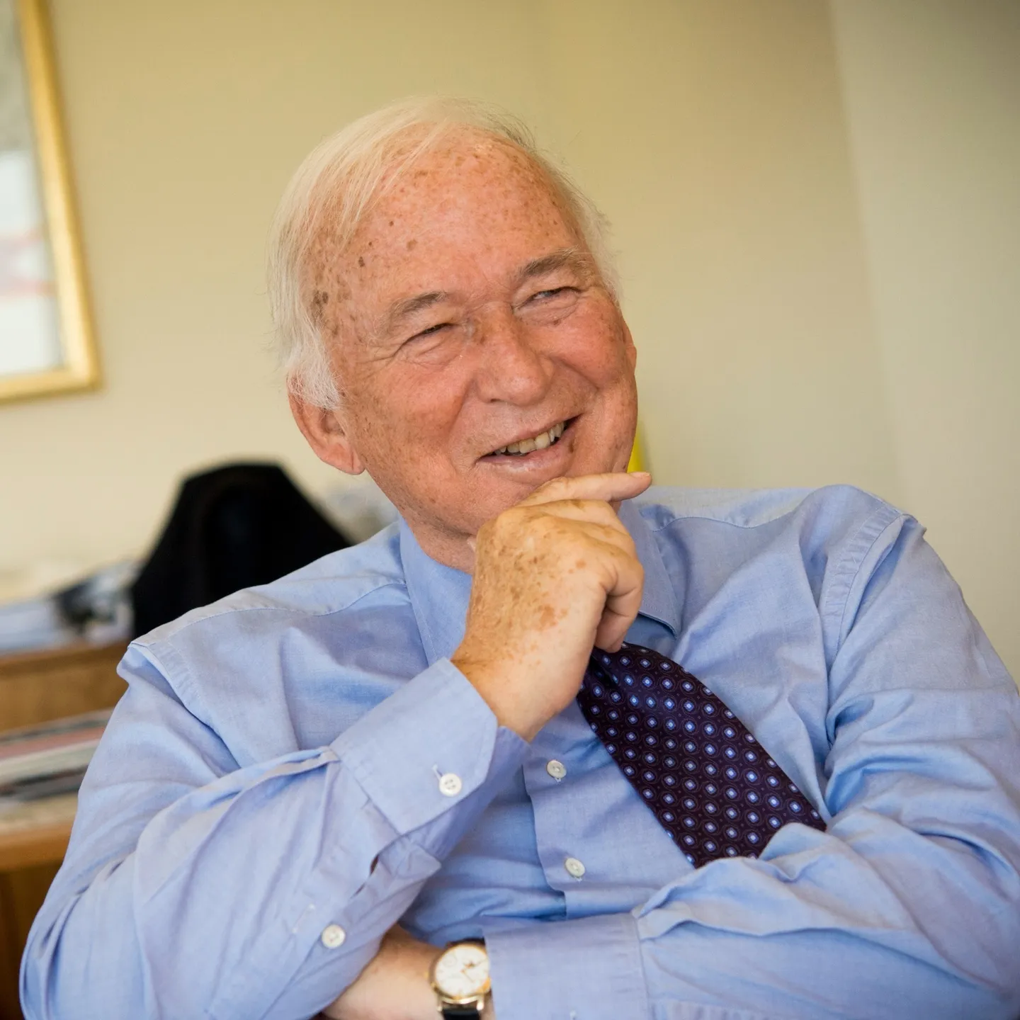 An older man in a blue shirt and tie with his hand at his chin, in his office