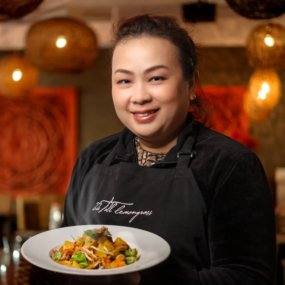 A chef in a black apron holding a plated dish in her restaurant