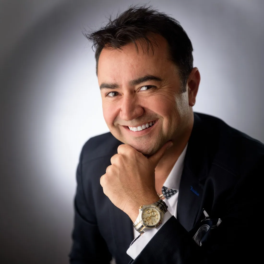 A smiling man in a dark jacket resting his chin on his hand
