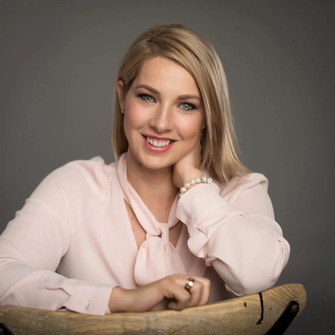 A woman in a pink blouse resting her chin on her hand as she leans on a chair