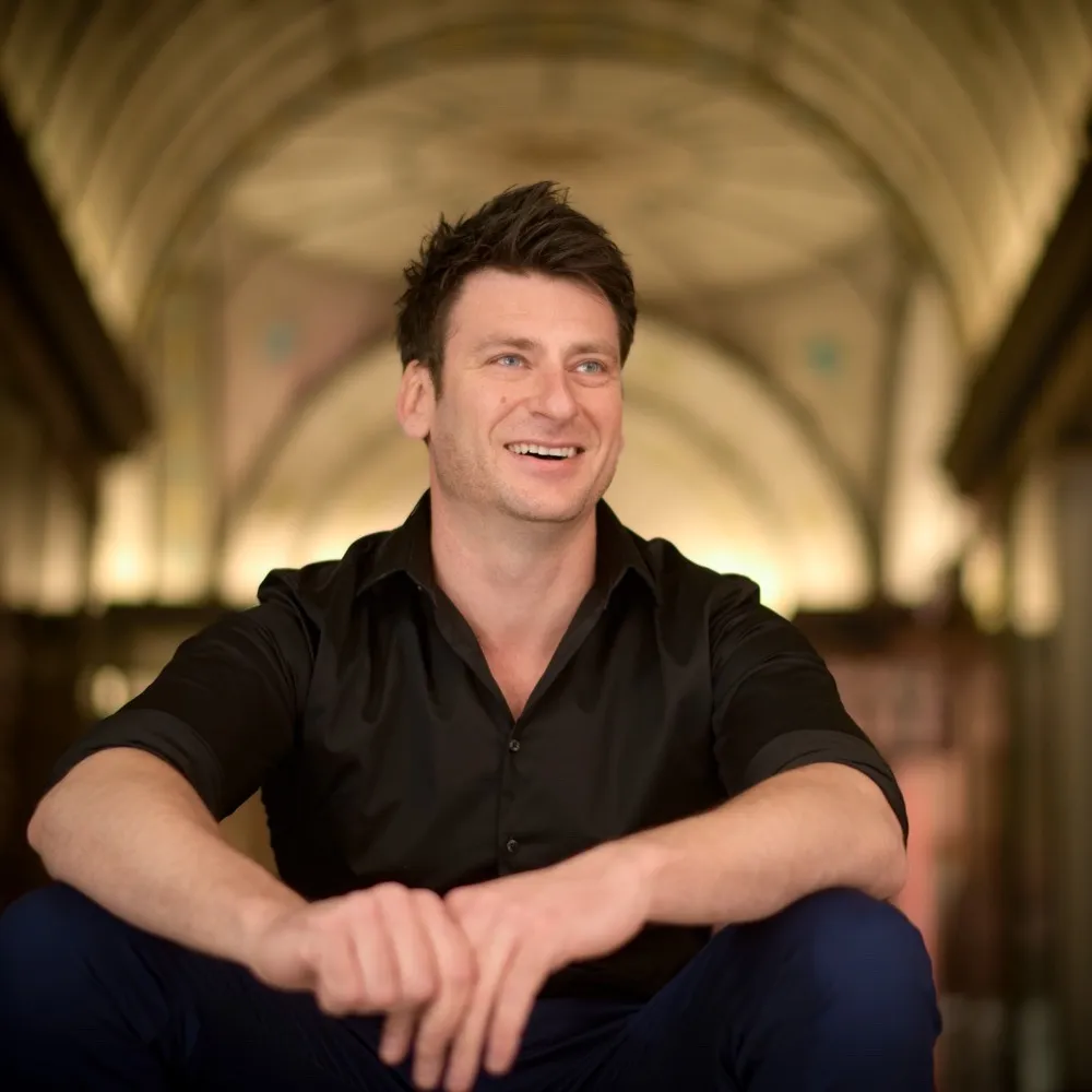 A smiling man in a black shirt seated beneath an arched ceiling